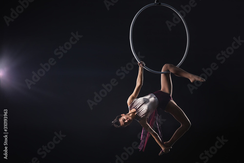 Young woman performing acrobatic element on aerial ring against dark background. Space for text photo