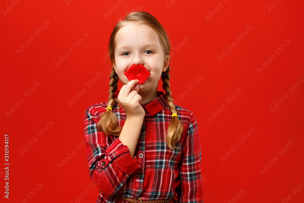 Funny cheerful little girl makes artificial fake lips on a red background. Child fooling around Funny cheerful little girl makes artificial fake lips on a red background. Child fooling around