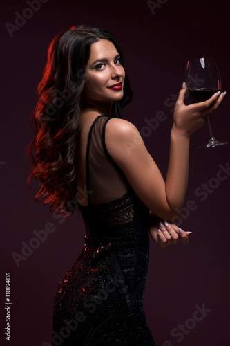 Glamour caucasian brunette woman in evening black dress on a neutral background. She pose sensually with glass of wine. Retro fashion mood.