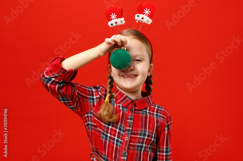Funny happy little girl with christmas ball decoration. A child in a red dress and mittens headband on a colored background