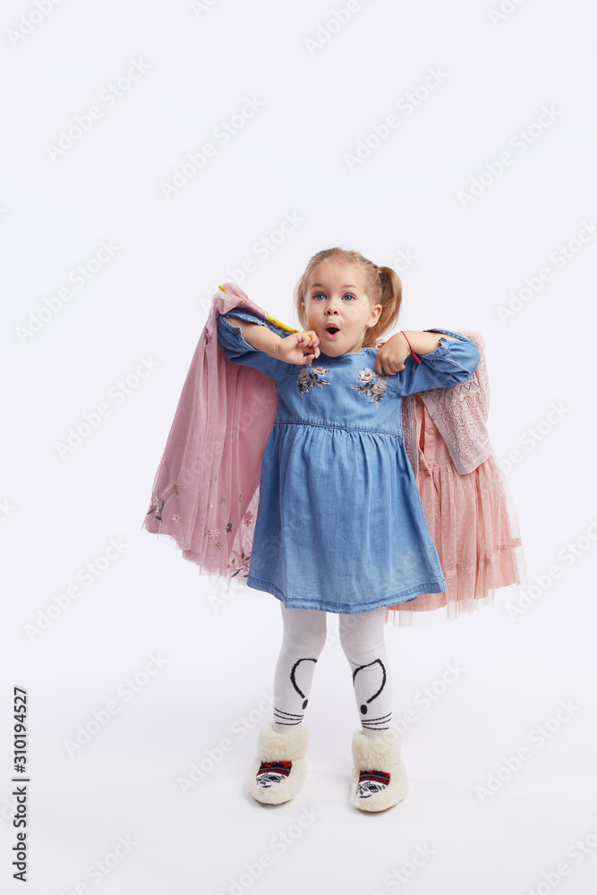 Little child girl chooses outfits for the holiday. Holds hangers with dresses in his hands. White studio background Little child girl chooses outfits for the holiday. Holds hangers with dresses in his hands. White studio background