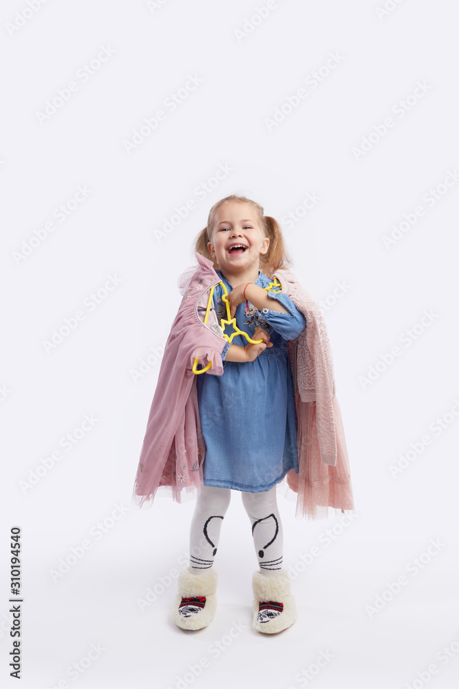Little child girl chooses outfits for the holiday. Holds hangers with dresses in his hands. White studio background Little child girl chooses outfits for the holiday. Holds hangers with dresses in his hands. White studio background