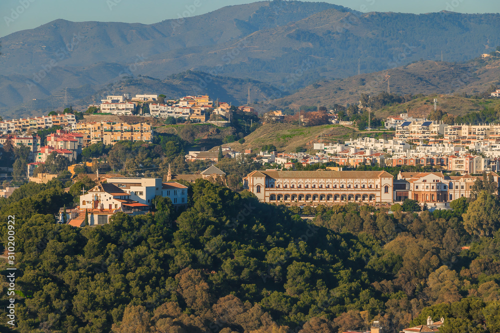 Panoramic view in Malaga from the hill of the Barcenillas district with Sagrada Familia El Monte. Hills with forest, trees, and apartment buildings on a sunny day. University with buildings