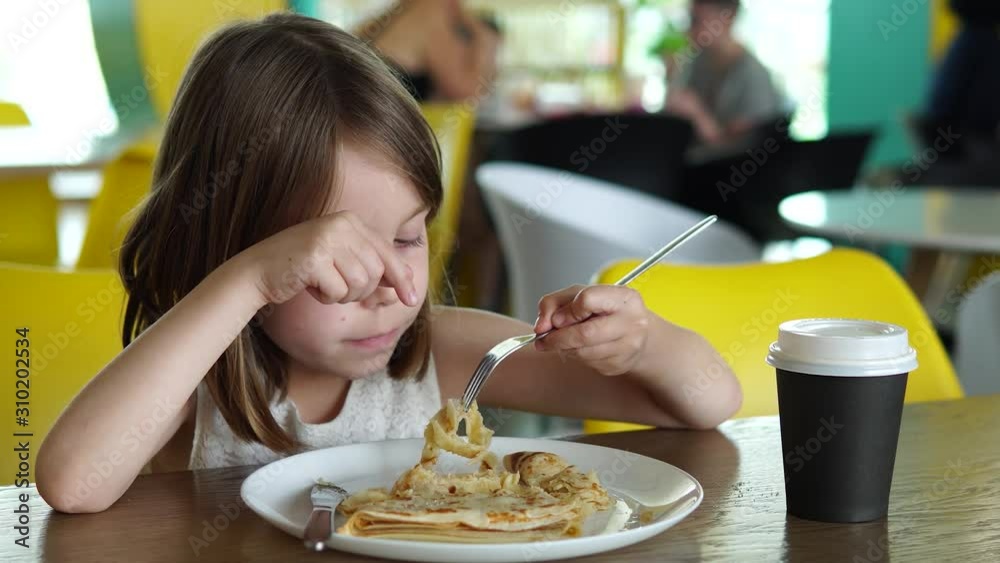 Close-up portrait of a girl of Russian appearance. Toddler in a cafe at the table, food court in a shopping or business center. In front of the child is a plate with a pancake. Close-up portrait of a girl of Russian appearance. Toddler in a cafe at the table, food court in a shopping or business center. In front of the child is a plate with a pancake.