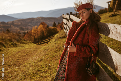 Outdoor fashion portrait of elegant, fashionable, confident woman wearing beret, red faux fur coat, holding small bag, posing in nature with mountains. Copy, empty space for text