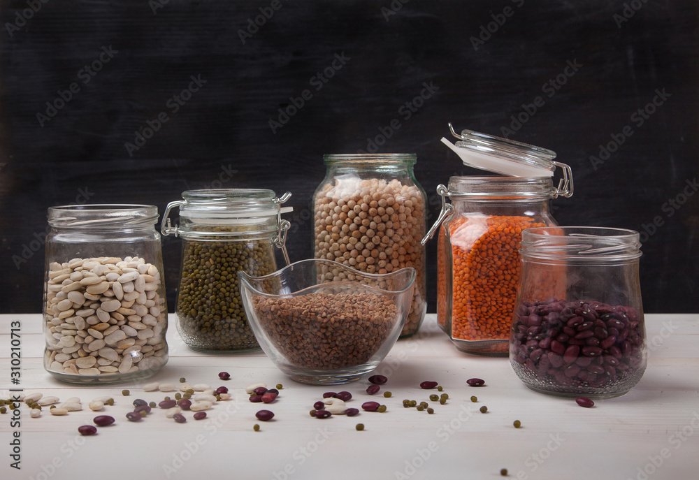 Red and white beans, peas, mung bean, red lentils in glass jars stand on a white wooden table on a black background Red and white beans, peas, mung bean, red lentils in glass jars stand on a white wooden table on a black background