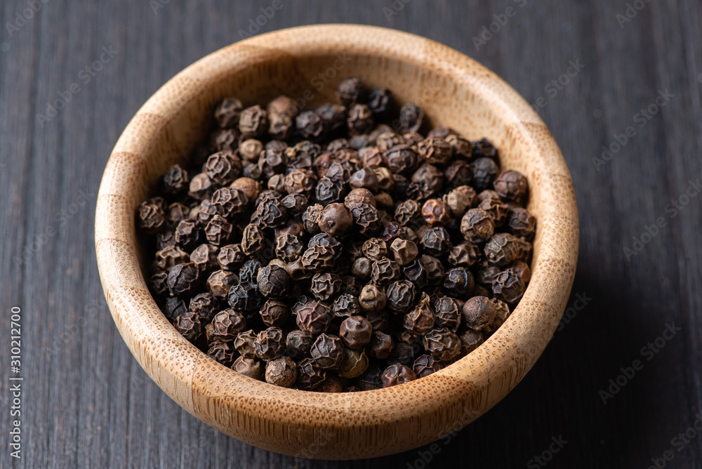 Whole black pepper in a small wooden bowl on a black wooden background. Macro, flat lay. Whole black pepper in a small wooden bowl on a black wooden background. Macro, flat lay.