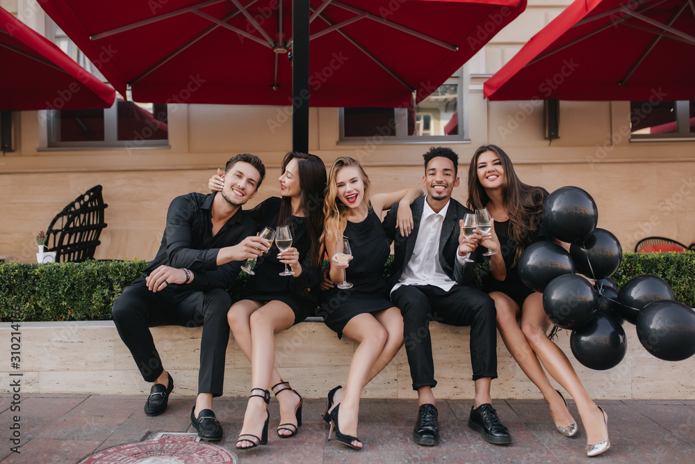 Fototapeta premium Brunette woman in elegant shoes embracing husband while he clink glasses with her. Outdoor portrait of loving couple celebrating anniversary with friends in favorite cafe.