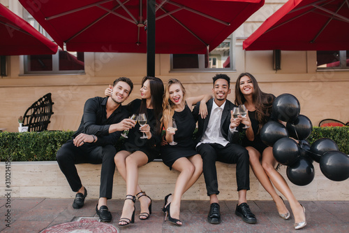 Brunette woman in elegant shoes embracing husband while he clink glasses with her. Outdoor portrait of loving couple celebrating anniversary with friends in favorite cafe.