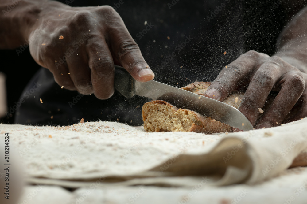 Close up of african-american man slices fresh cereal, white bread, bran with a kitchen knife on wooden table. Healthy eating, nutrition, craft product. Gluten-free food, vegan lifestyle, organic taste
