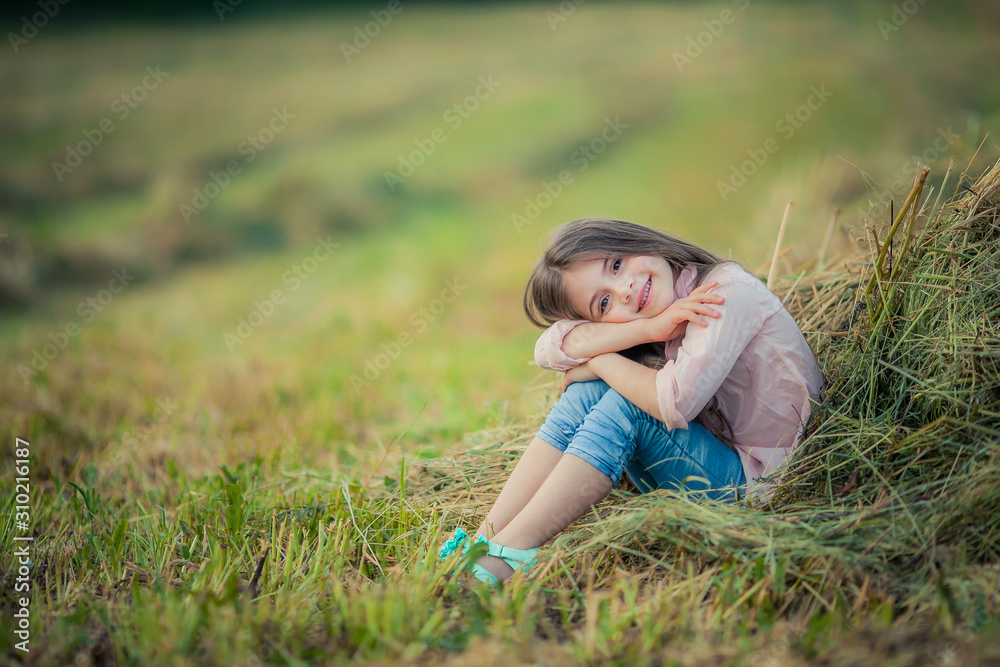 Fototapeta premium A girl sits in a field near a pile of freshly mown green hay.