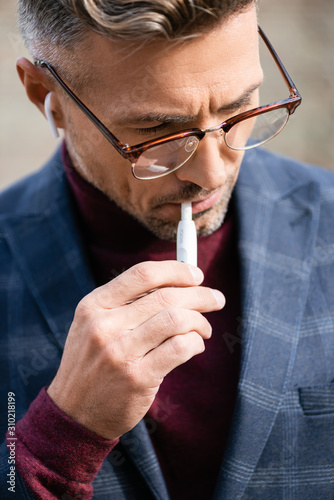 KYIV, UKRAINE - OCTOBER 11, 2019: Stylish businessman in eyeglasses and wireless earphones smoking iqos photo