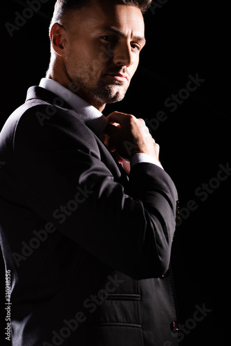 Handsome businessman adjusting shirt collar while looking at camera isolated on black
