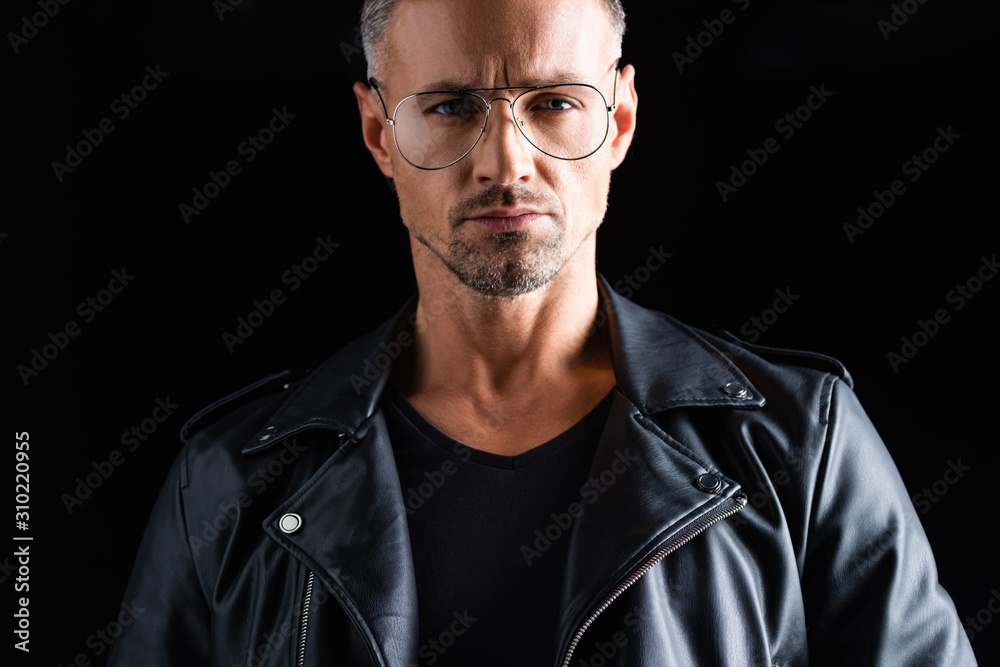 Confident stylish man in sunglasses and leather jacket looking at camera isolated on black