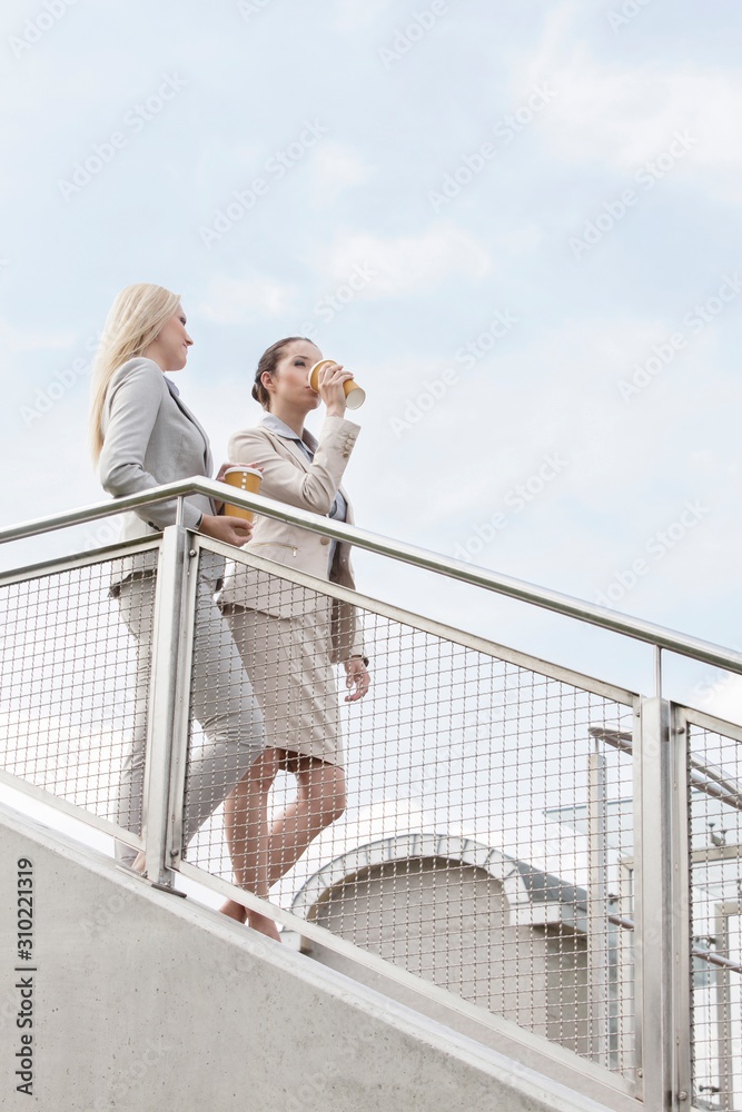 Young businesswomen with disposable coffee cups moving down stairs against sky