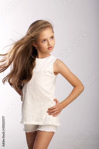 young beautiful blonde with wavy hair in a white summer dress smiles and poses in the studio at the camera on a gray background with blue eyes with a confident look looks into the distance