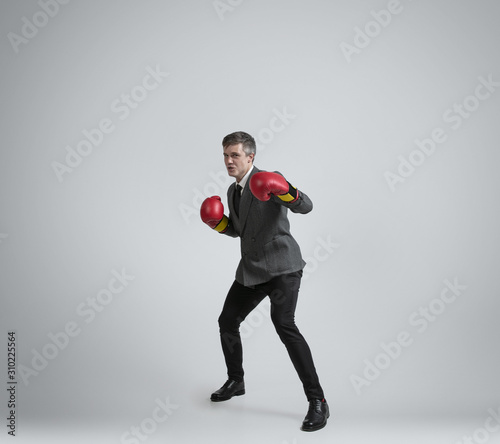 Wild and young. Caucasian man in office clothes boxing with two red gloves on grey background. Businessman training in motion, action. Unusual look for sportsman, activity. Sport, healthy lifestyle. © master1305