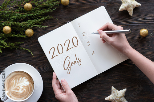 Female hands write 2020 new year resolution goal wish list in notebook on brown wooden table background, woman make notes plan for better changes self improvement motivaion concept, flat lay top view photo