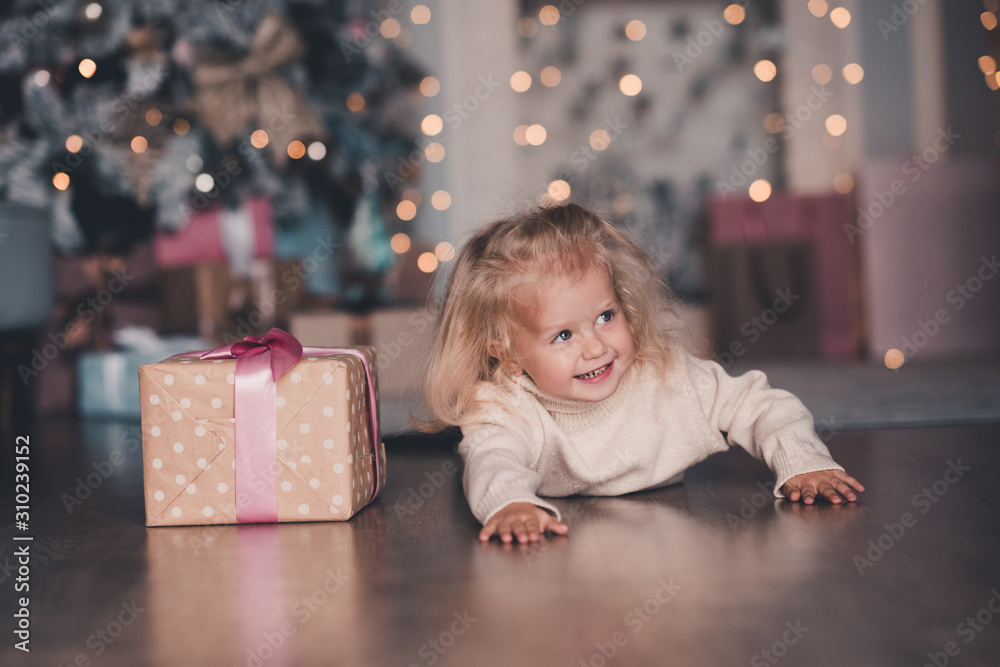 Happy child girl 4-5 year old crawling on floor with Christmas present over xmas tree with glowing lights closeup. New Year. Childhood. Happy child girl 4-5 year old crawling on floor with Christmas present over xmas tree with glowing lights closeup. New Year. Childhood.