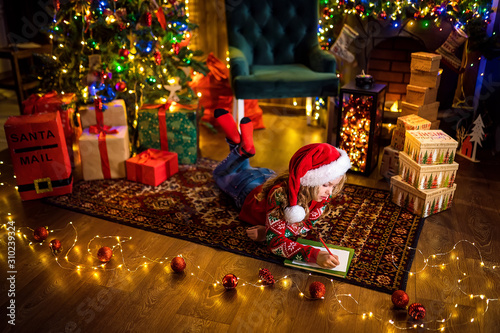 Little charming girl in Santa hat is lying on the floor and writes a letter, draws with a pencil. lights of garlands. warm comfortable room