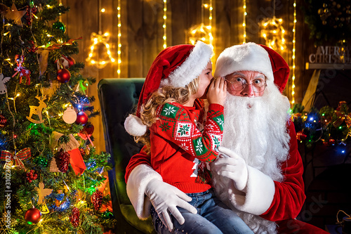 Little girl whispering in Santa's ear. Telling a secret. Revealing the gift you would like to win. Talking about how she behaved during the year. photo