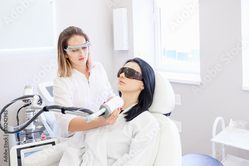Charming female beautician does ultrasonic face cleaning pretty woman client sitting in a chair in a white coat and safety glasses in beauty spa clinic.  oncept of skin cleansing procedures