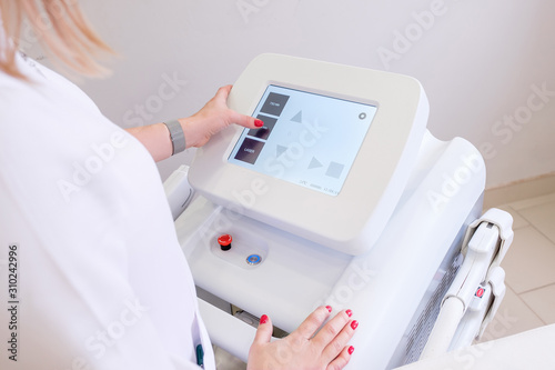 Rear view of a female beautician in white coat presses on display of device to select desired procedure. Concept of modern high-tech equipment for anti-aging procedures. medical examination of patient