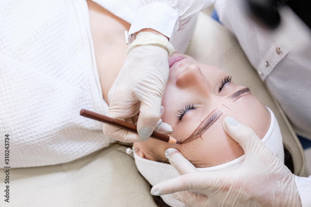 Fototapeta premium Close-up of the hand of a beauty master woman sketching a new eyebrow shape to a young female client. Concept of eyebrow tattoo and microblading