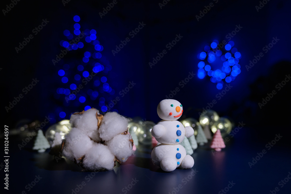 Fototapeta premium A sweet snowman from a pastry mastic on a background of marshmallow trees and a white mug with cocoa. Sweets and a bud of hlok in the dark against the background of a Christmas tree in the lights.
