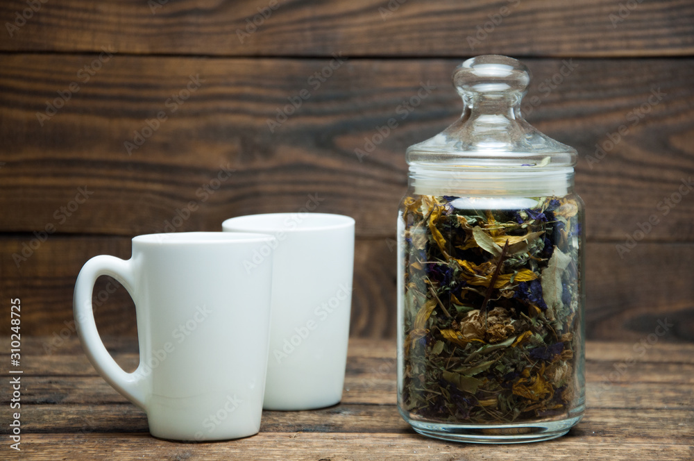 A white cup and a glass jar with herbal aromatic tea stands on a wooden table. Tea and coffee in the morning.