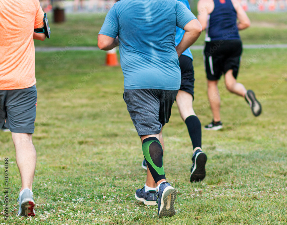 Rear view of sweaty runner with a compression calf sleeve racing a 10K on grass at a park Rear view of sweaty runner with a compression calf sleeve racing a 10K on grass at a park
