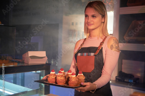 Young attractive woman is holding a tray with freshly prepared cakes at bakery store.