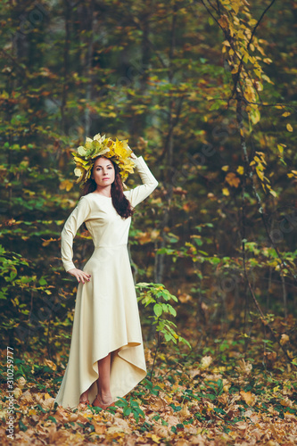portrait of a brunette in full growth in the autumn forest in a light dress in a wreath of leaves © Nikita