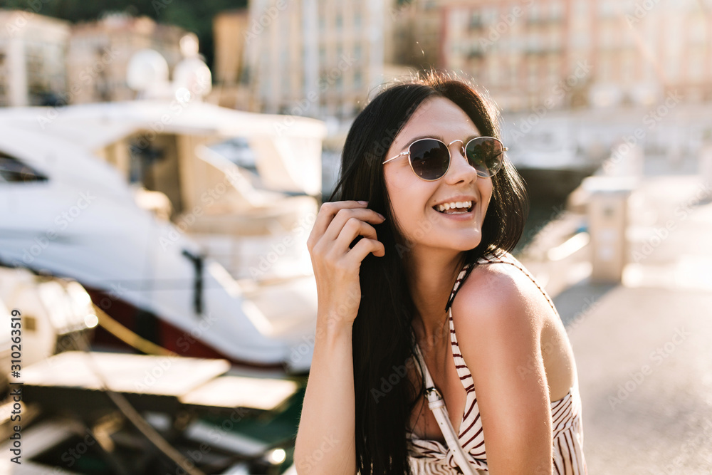 Excited girl with long black hair happy laughing while sitting outdoor with boats on background. Portrait of blissful smiling brunette woman in sunglasses having fun on city pier in sunny day.