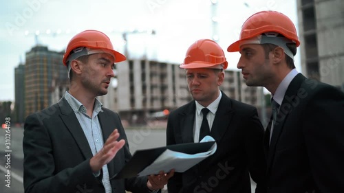 4k. Ultra HD. Three Young successful architects in orange helmets and suits with blueprint discussing sketch of new building construction on text pad. Static middle shot with crane and beams bg. photo