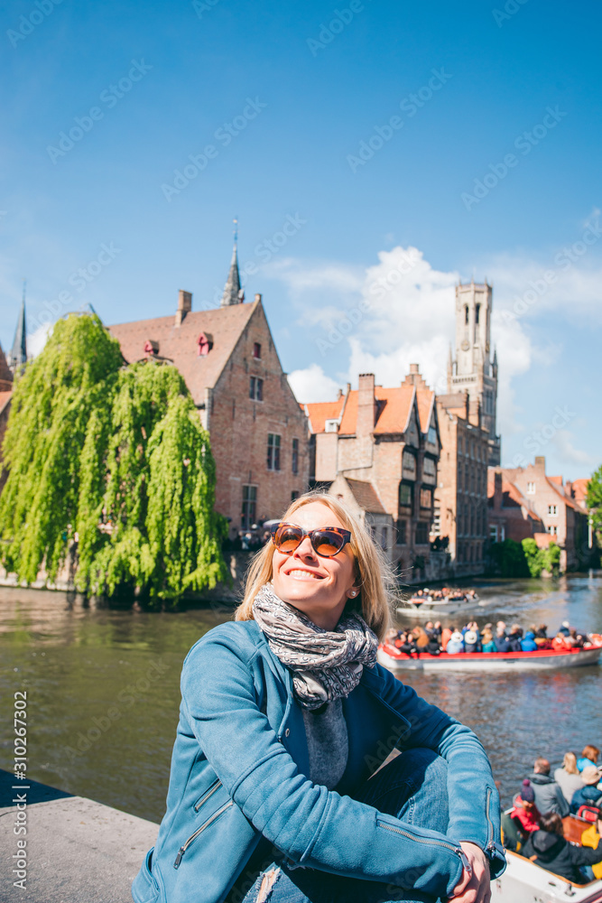 Fototapeta premium A beautiful young girl sits on the background of a famous tourist spot with a canal in Bruges, Belgium