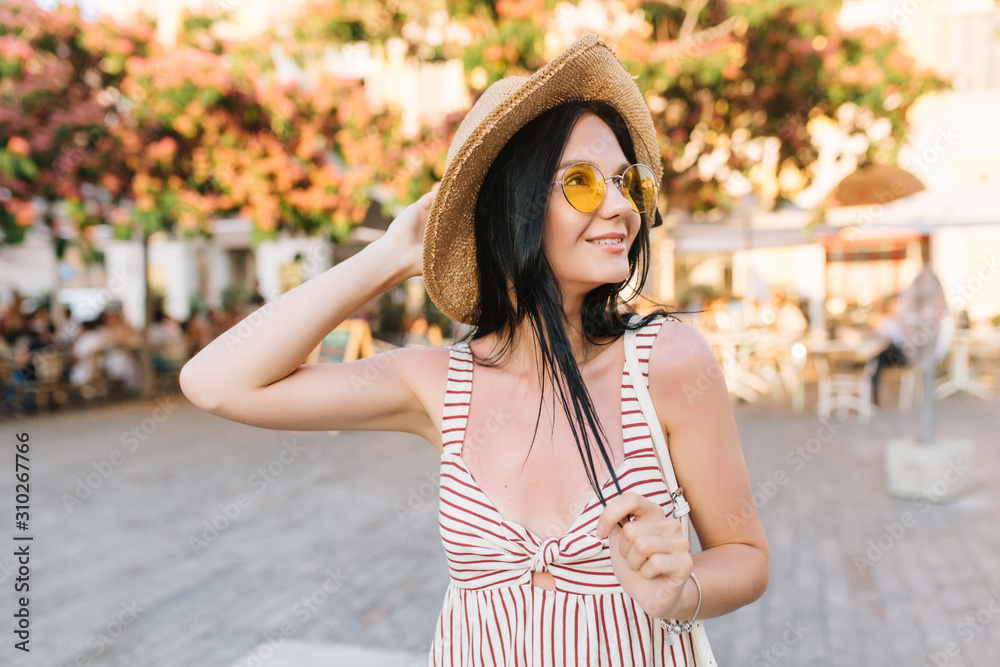 Inspired graceful girl wearing trendy glasses and summer hat spending time outdoor, enjoying warm day. Cute tanned woman in stylish accessories going to beach and looking around with interest.
