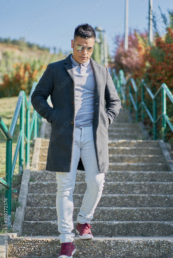 portrait of a handsome Hispanic boy standing on the stairs with good pants and urban clothes: jeans, blue shirt and dark jacket and sunglasses on his face portrait of a handsome Hispanic boy standing on the stairs with good pants and urban clothes: jeans, blue shirt and dark jacket and sunglasses on his face