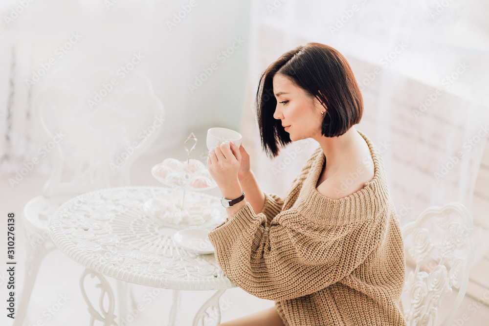 Charming young woman in beige sweater enjoy her coffee sitting at the coffee table Charming young woman in beige sweater enjoy her coffee sitting at the coffee table