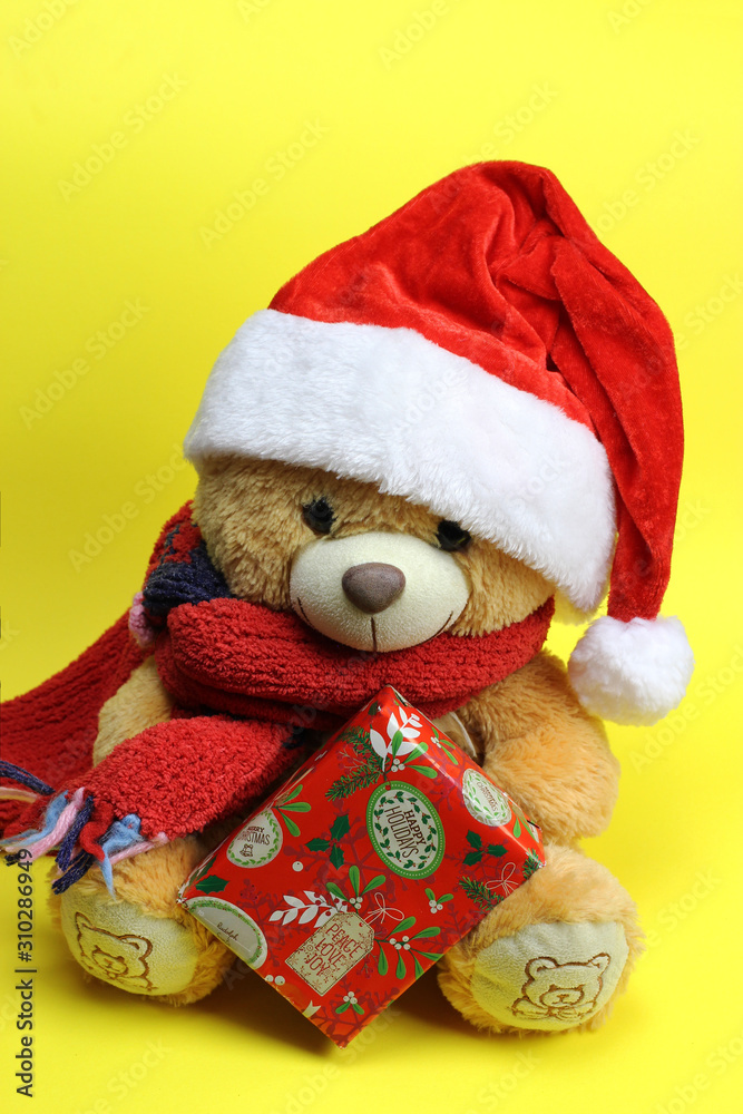 Teddy bear in Santa Claus hat holding a Christmas gift. yellow background