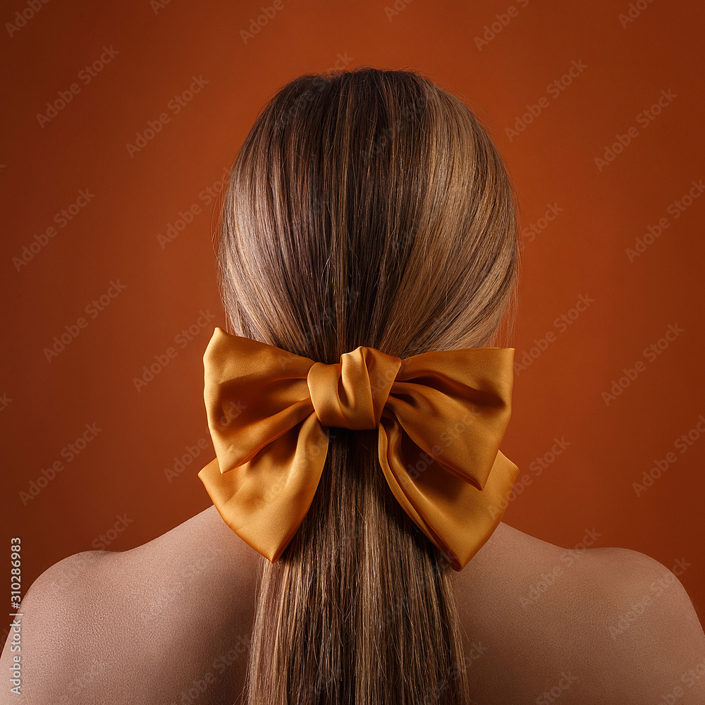 blond female hairstyle with color ribbon on orange wall background. close  up of blonde girl head with carroty bow in her hair, rear view. fashion  style photoshoot of woman in studio Stock