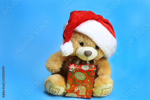 Teddy bear in Santa Claus hat holding a Christmas gift. Blue background