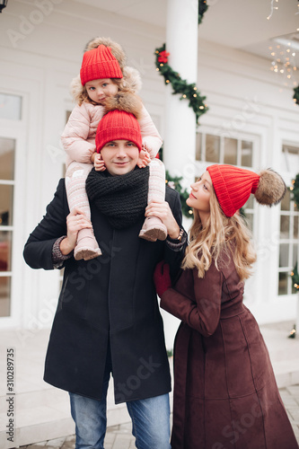 Stock photo of attractive happy Caucasian family of three in red hats in the street. Pregnant mother is looking at her husband with love. Little daughter is sitting on father s neck. They are smiling photo