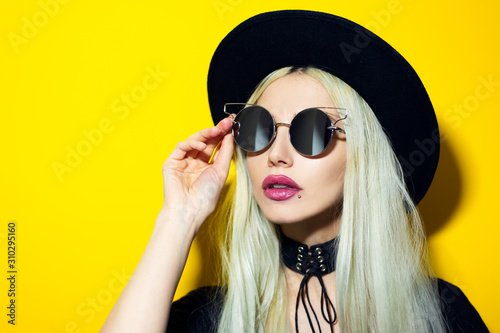 Close-up studio portrait of young blonde girl wearing black sunglasses and hat on yellow background. © Lalandrew
