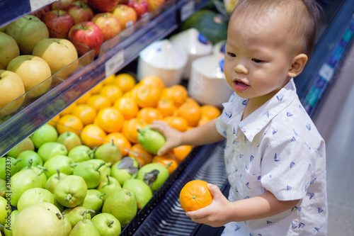 Cute little Asian 18 months / 1 year old toddler baby boy child shopping in a supermarket. Kid standing choosing and holding fruit in grocery store, Kid first experience concept - Selective focus photo