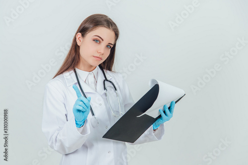 Young female intern standing in medical uniform  holding clipboard  checking data  giving finger up  calling to pay attention.