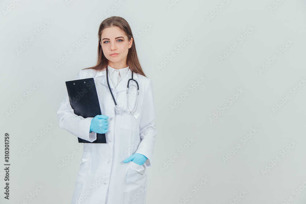 Happy young female intern standing in medical uniform, holding clipboard, putting hand into the pocket of medical coat. Happy young female intern standing in medical uniform, holding clipboard, putting hand into the pocket of medical coat.