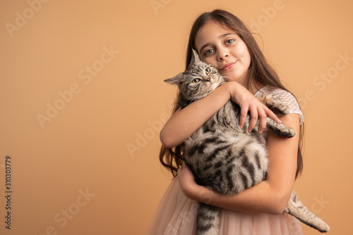 Beautiful cute brunette girl with loose hair, blue eyes and healthy skin hugging her grey cat and looking at the camera isolated over orange background