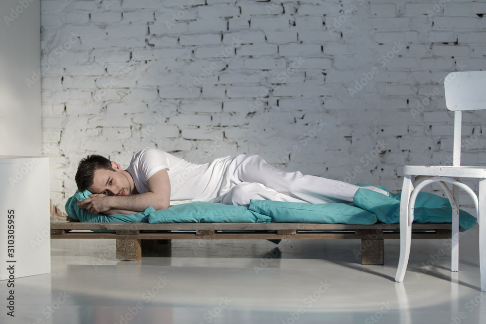 Fototapeta premium Young guy in white clothes depressed on a bed in a white room