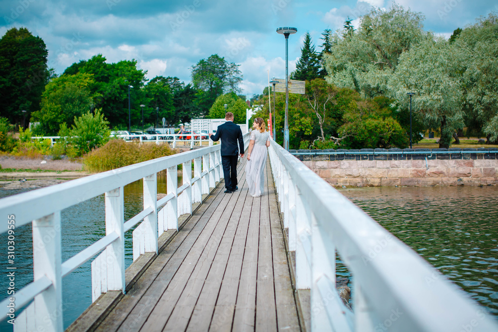 The bride and groom walk along the painted light wooden pier on a cloudy summer day, holding hands. The bride is pregnant. Helsinki. Finland